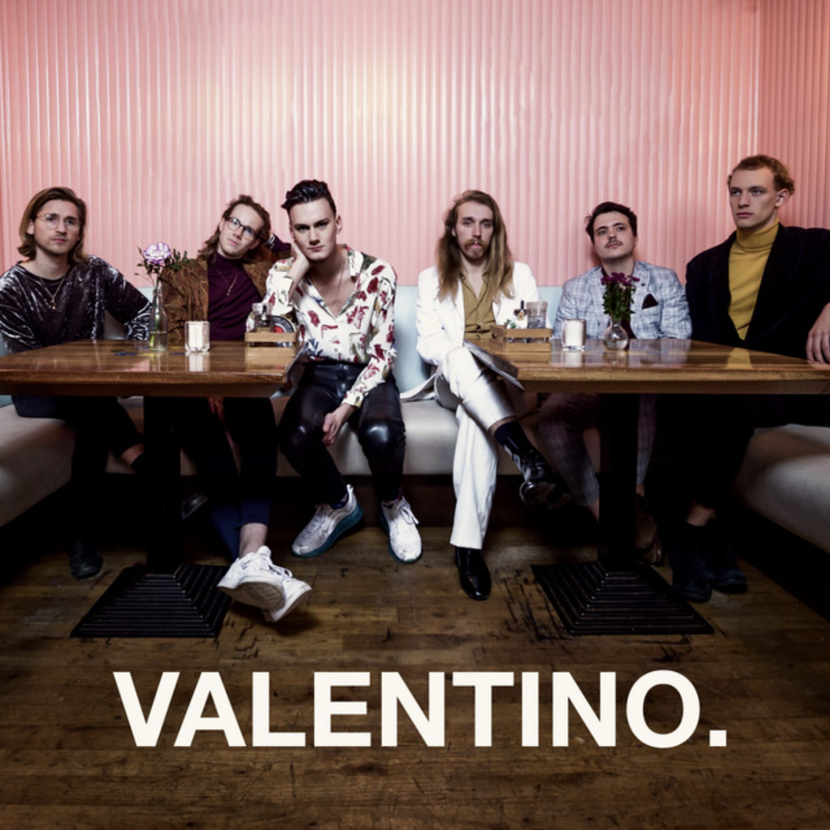 Band portrait of six musicians seated around a table against a pink striped wall.