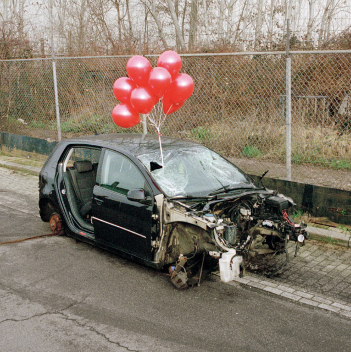 Damaged black car parked beside a fence with bright red balloons floating above it.