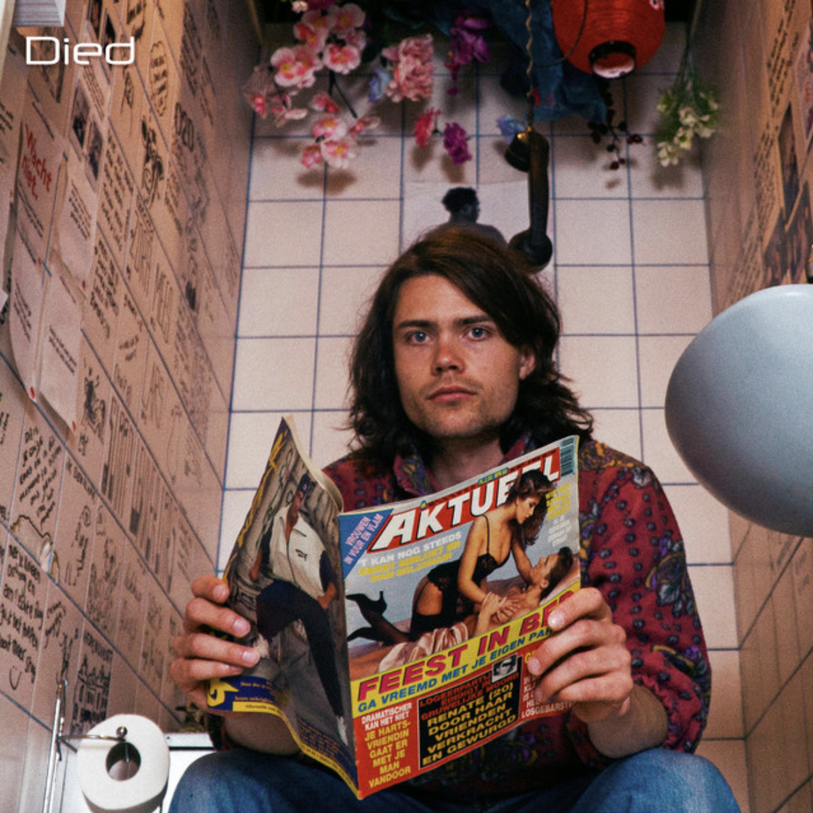 Portrait of a long-haired person holding a magazine while seated in a room decorated with balloons and flowers.