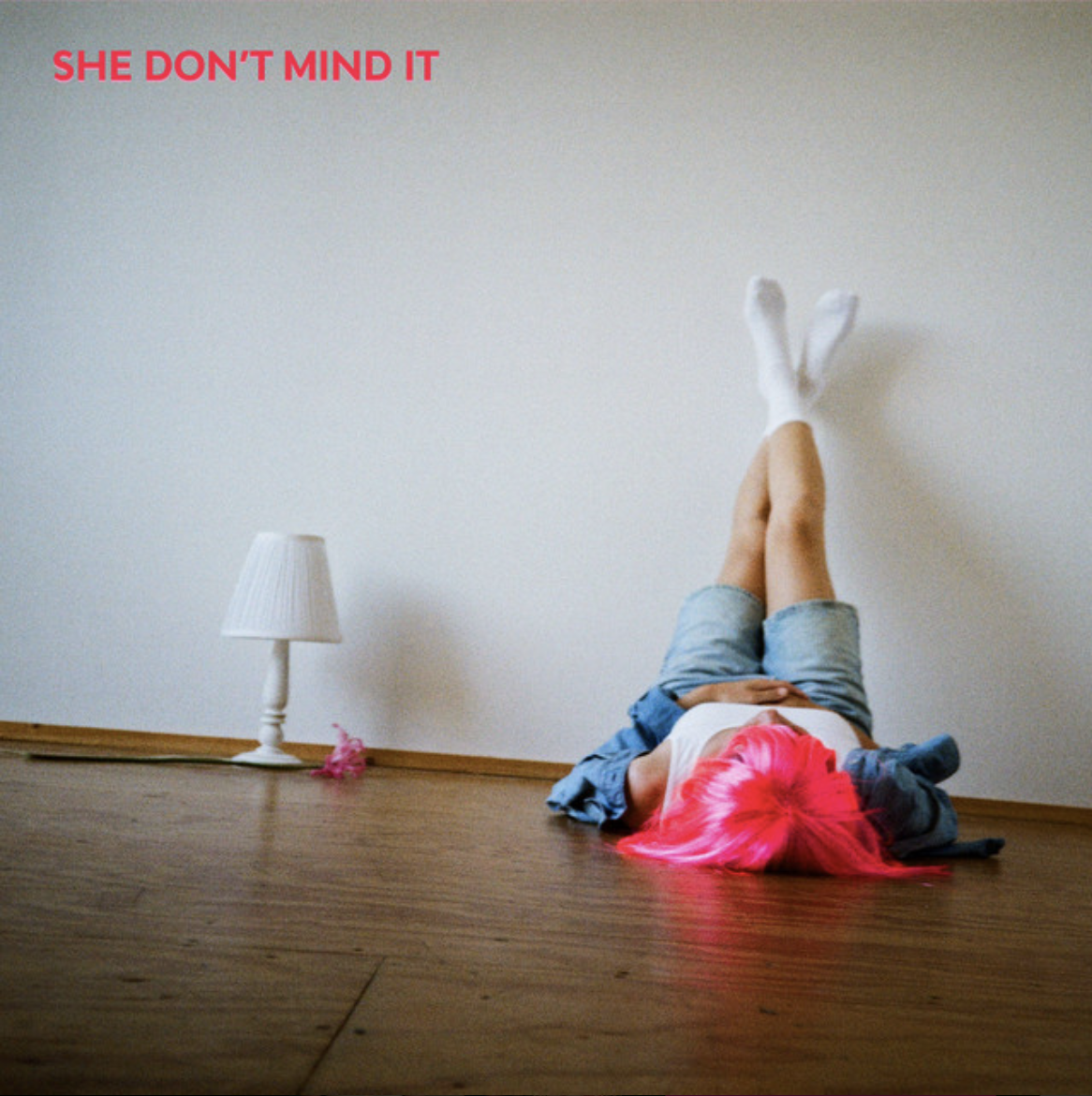 Person with bright pink hair lying upside down on a wooden floor with legs raised against a minimalist white wall.