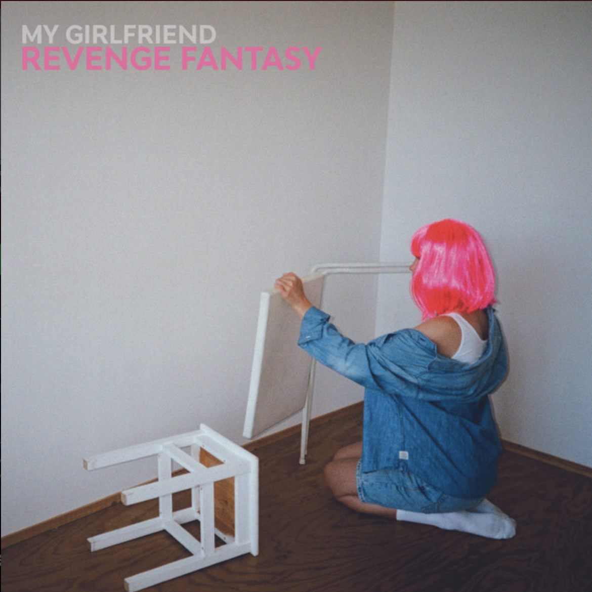Person wearing a bright pink wig sitting on the floor beside an overturned white chair in a sparse room.